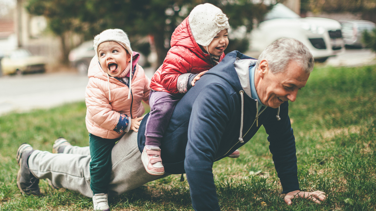 A photo of a playful grandfather and granddaughter. They are casually dressed and playing in the park. They exercise together. A grandfather is exercising while granddaughters are sitting on his back.