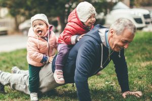 A photo of a playful grandfather and granddaughter. They are casually dressed and playing in the park. They exercise together. A grandfather is exercising while granddaughters are sitting on his back.